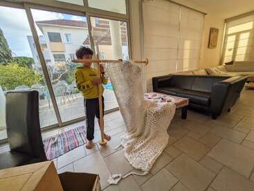 A young boy stands barefoot by a large window, working on a wooden loom with white yarn in a cozy, sunlit living room with modern furniture.