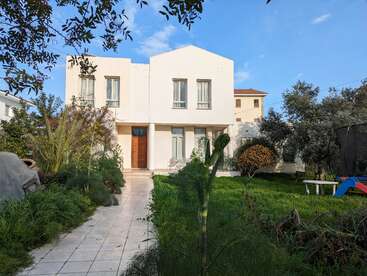 A modern white two-story house with a flat roof stands amidst a lush green garden, featuring trees, shrubs, a trampoline, pathway, and clear blue sky.