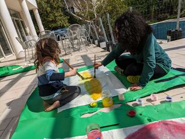 A child and an adult sit outdoors on a patio, painting a large sheet with yellow paint, surrounded by jars and containers of colorful paint.
