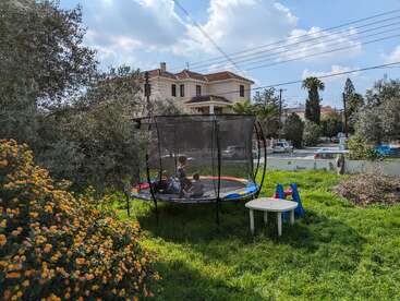 Two children play inside a trampoline in a grassy backyard. Flowers bloom nearby, a table and chairs sit outside, and houses stand in the background under blue skies.