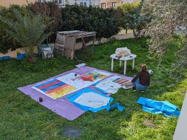 A woman is painting colorful abstract art on large canvases spread on grass in a backyard, surrounded by trees, art supplies, and a small table.