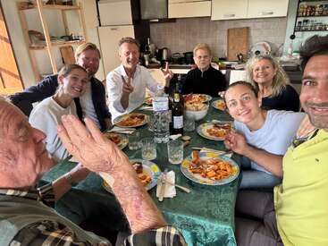 Un grupo de ocho personas sentadas alrededor de una mesa disfrutan de una comida juntos. Sonríen, parecen felices y relajadas en un acogedor entorno de cocina.