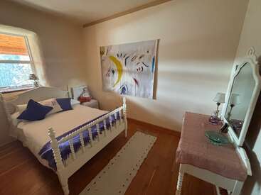 This cozy bedroom features a white vintage bed, abstract wall art, purple accents, a window, a side table, a vanity with mirror, and wooden floors.