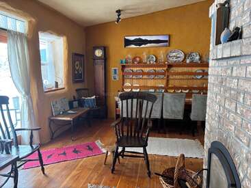 This cozy room features wooden floors, a brick fireplace, rocking chairs, a grandfather clock, large window, a shelf with pottery, and a dining table.
