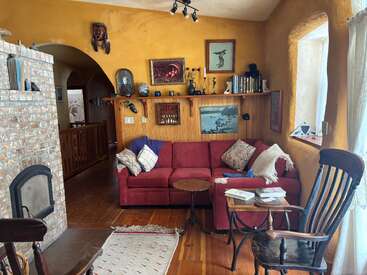A cozy living room with a red sectional sofa, wooden furniture, bookshelf, art, warm yellow walls, brick fireplace, decorative objects, and sunlight streaming through curtains.