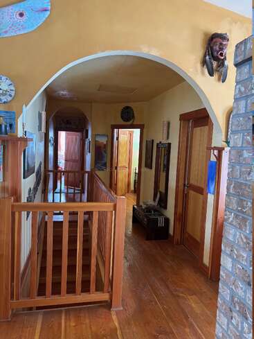 A cozy hallway with wooden floors and doors, arched ceiling, eclectic decorations—including a fish sculpture and a mask—and a staircase with wooden railing. Warm lighting.
