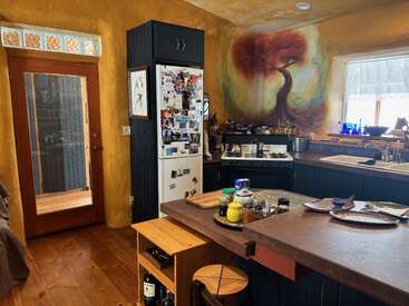 This cozy kitchen features warm yellow walls, a tree mural, wooden floors, dark cabinets, a cluttered fridge, countertop items, a door, window, and natural light.