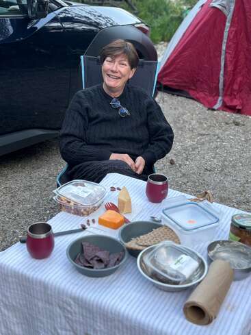 A woman sits smiling at a picnic table outdoors, with snacks, cheese, and crackers. Behind her, a car and a red tent are visible. Relaxed atmosphere.