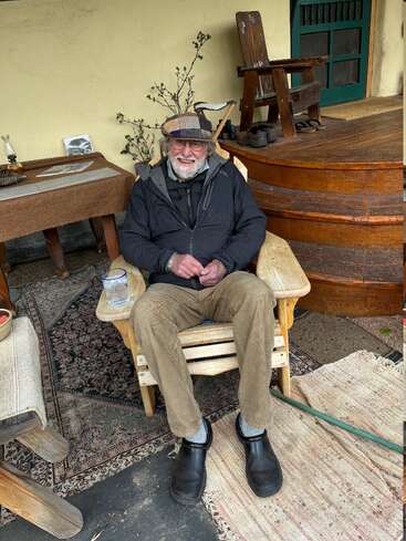 An elderly man with a gray beard and glasses smiles, sitting in a wooden chair on a porch. He wears a hat, jacket, and brown pants.