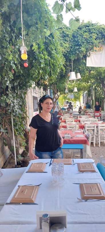 Une femme se tient près d'une table dans un restaurant en plein air, entourée de verdure, de nappes rouges et de chaises blanches, créant une atmosphère paisible et accueillante.