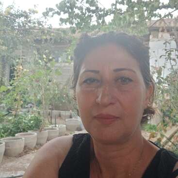 Une femme aux cheveux noirs et aux sourcils arqués pose pour un selfie en plein air. Derrière elle, des plantes en pot, du feuillage vert et un bâtiment rustique.