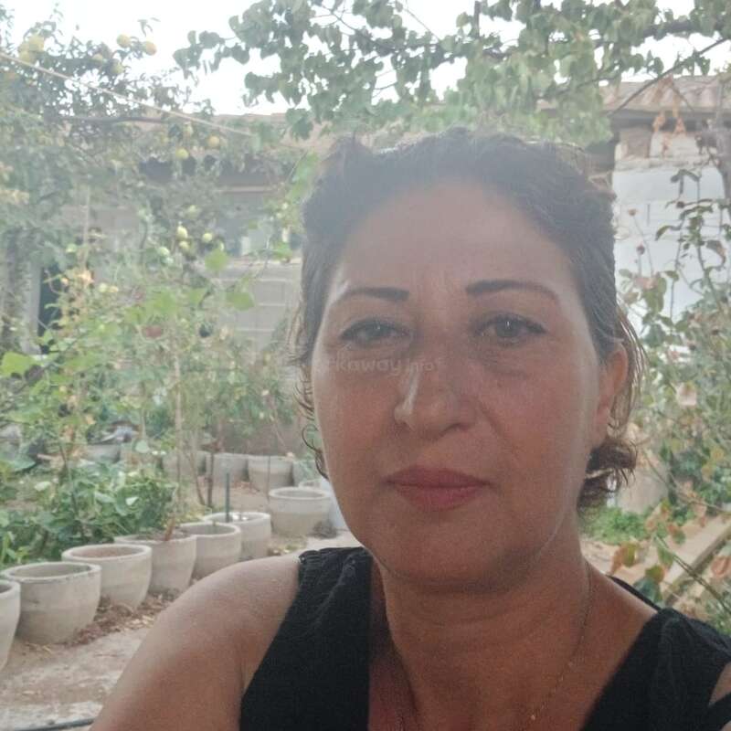 Une femme aux cheveux noirs et aux sourcils arqués pose pour un selfie en plein air. Derrière elle, des plantes en pot, du feuillage vert et un bâtiment rustique.