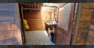 This image shows a small wooden kitchen with a corrugated roof, gravel floor, a counter with a stove, some shelves, a door, and a yellow raincoat.