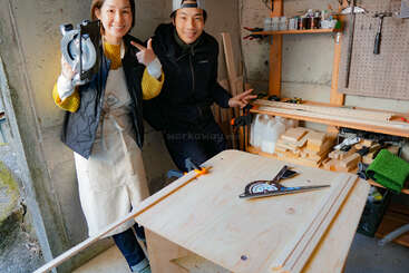 Deux personnes sourient dans un atelier de menuiserie. L'une d'elles tient une scie circulaire. On aperçoit des outils, des morceaux de bois et un établi. Toutes deux ont l'air joyeuses et enthousiastes.