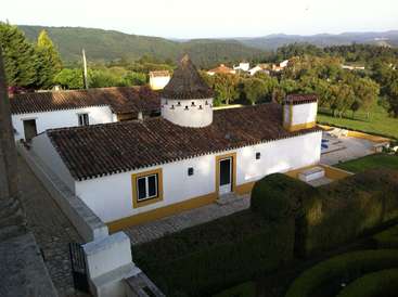 The image depicts a white house with a brown tiled roof, featuring a tower and chimney, set against a backdrop of rolling hills and trees.