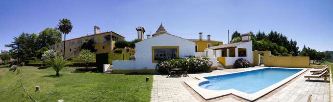 The image depicts a large, multi-story yellow and white house with a pool and patio area, surrounded by a well-manicured lawn and trees.