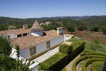 The image depicts a white house with a tiled roof and a garden, set against a backdrop of rolling hills and mountains under a clear blue sky.