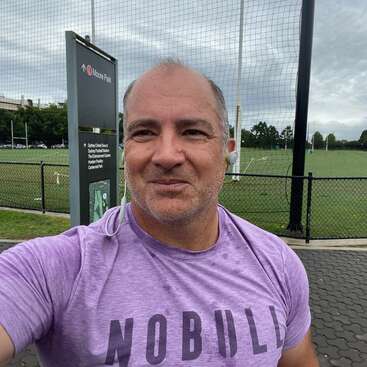 A imagem mostra um homem tirando uma selfie em frente a um campo de futebol, usando uma camiseta roxa com a palavra \"NOBULL\" e fones de ouvido.