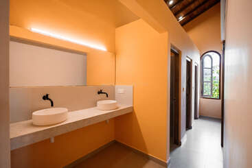 Esta imagen muestra un cuarto de baño moderno con paredes de color melocotón, dos lavabos redondos, grifos negros, un espejo grande y un pasillo con puertas y una ventana en arco.
