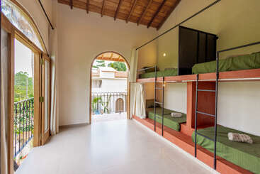 Esta imagen muestra una habitación compartida luminosa y espaciosa con cuatro literas, ropa de cama verde, grandes ventanas, techo de madera y vistas al exterior a través de puertas arqueadas.