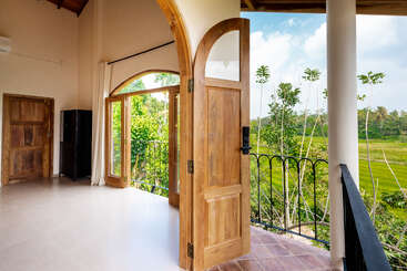 Una habitación soleada con puertas de madera se abre a un balcón con vistas a exuberantes campos verdes y altos árboles bajo un cielo azul brillante. Interior tranquilo, acogedor y espacioso.