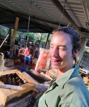 A woman in a green shirt stands in a rustic outdoor workspace, squinting and smiling. She is near a wooden box with a syringe on top.