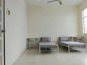 L'image montre une chambre simple et épurée avec deux lits simples, deux bureaux avec des chaises, un ventilateur de plafond, des murs blancs et une grande fenêtre.
