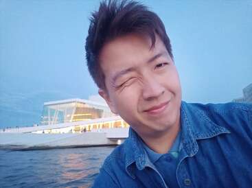 Un jeune homme vêtu d'une chemise en jean fait un clin d'œil à la caméra, souriant au bord de l'eau. Un bâtiment moderne en verre illuminé par les lumières du soir brille en arrière-plan.