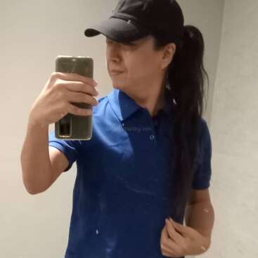 A person wearing a black cap and blue polo shirt takes a mirror selfie, holding their phone in one hand and their long hair in the other.