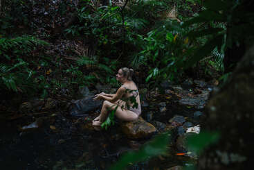 Uma mulher senta-se em uma pedra em um riacho raso da floresta, cercada por uma folhagem verde exuberante, desfrutando do ambiente natural e pacífico e da atmosfera tranquila e isolada.