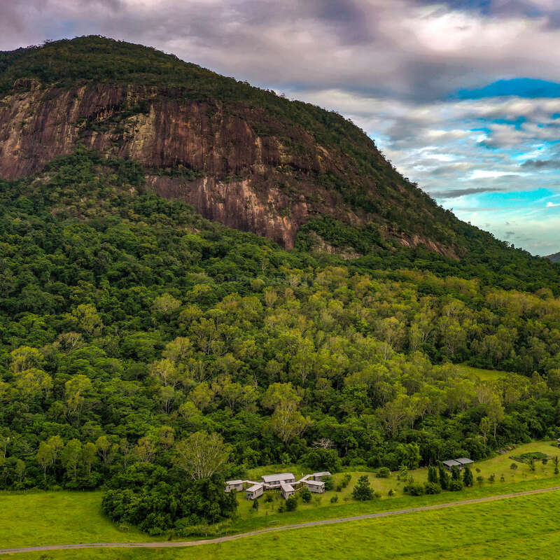 Uma floresta verde exuberante e uma planície gramada ficam no sopé de uma grande colina rochosa. Pequenos edifícios espalhados aninham-se pacificamente sob um céu vibrante e nublado.