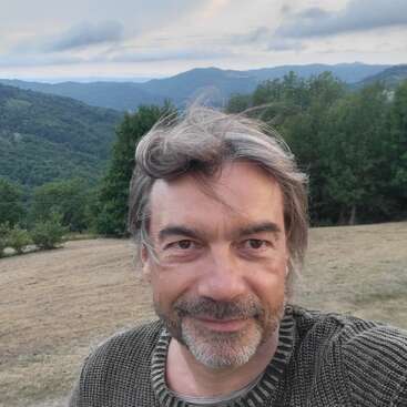 Un homme souriant aux cheveux gris et à la barbe prend un selfie en plein air. Derrière lui, des collines verdoyantes, des arbres et un ciel nuageux au crépuscule.