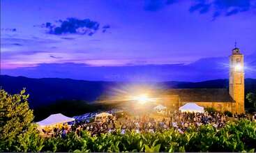 A vibrant outdoor evening event with crowds, white tents, and a glowing light near a rustic stone building, set against a mountainous landscape at sunset.