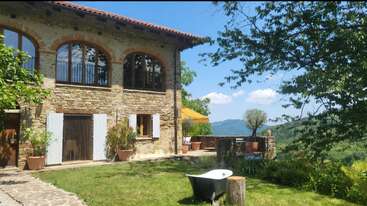 A rustic stone house with arched windows sits amid lush greenery. A bathtub and stump rest on the lawn. Mountain views and clear blue sky surround the scene.