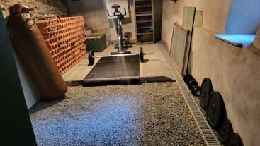 This image shows a home gym with a punching bag, exercise bike, dumbbells, weight plates, mat, and rustic walls. The floor is covered with gravel.
