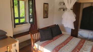 This cozy bedroom features a wooden bed with patterned bedding, two dark pillows, an open window with a garden view, and a hanging mosquito net.