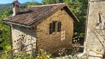 A charming small stone cottage with a tiled roof sits surrounded by greenery and sunlight. Wooden shutters, a rustic chimney, and peaceful rural atmosphere abound.