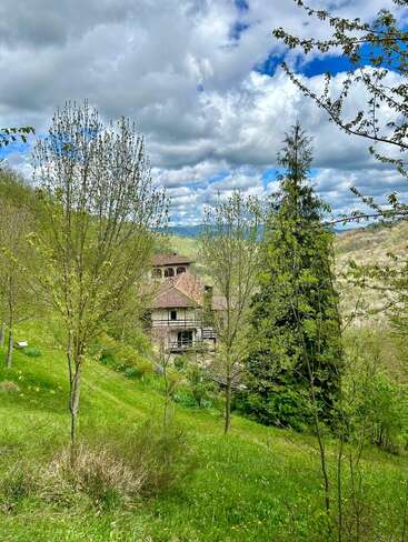 A charming countryside house nestles among fresh green trees and rolling hills, beneath a dramatic cloudy sky, radiating tranquility and natural beauty in springtime.