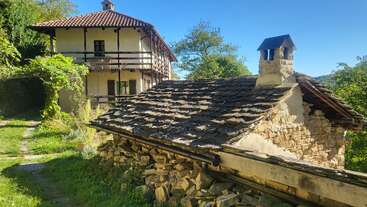 A rustic scene with two houses: one old stone cottage with a shingled roof and another larger, wooden balcony home, surrounded by greenery and bright sunlight.