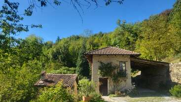 A rustic stone house with a tiled roof sits peacefully amidst lush green trees under a bright blue sky, surrounded by nature and serene countryside charm.