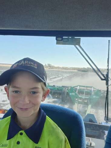 Das Bild zeigt einen Jungen, der eine Mütze und ein Warnwestenhemd trägt und in der Kabine eines Traktors sitzt, durch dessen Windschutzscheibe ein Feld und landwirtschaftliche Geräte zu sehen sind.