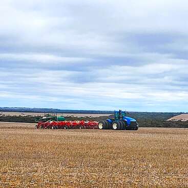 Das Bild zeigt einen blauen Traktor auf einem Feld, der ein rotes landwirtschaftliches Gerät zieht, vor dem Hintergrund eines bewölkten Himmels und sanfter Hügel in der Ferne.