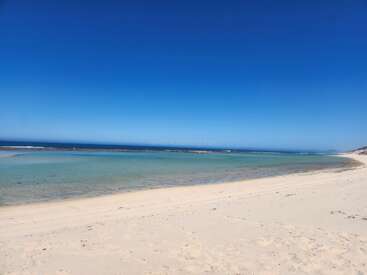 Das Bild zeigt eine heitere Strandszene mit einer weiten Sandfläche, einem ruhigen Gewässer und einem klaren blauen Himmel, der ein Gefühl der Ruhe hervorruft.