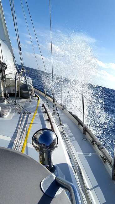 Das Bild zeigt ein Segelboot, das durch den Ozean fährt, mit Wellen, die gegen die Bordwand schlagen, und einem klaren blauen Himmel darüber, der ein Gefühl von Abenteuer und Freiheit hervorruft.