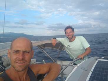Das Bild zeigt zwei Männer auf einem Boot im Meer, wobei ein Mann steht und der andere sitzt und beide in die Kamera lächeln.