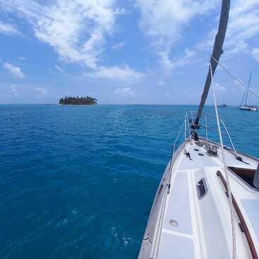 Das Bild zeigt eine ruhige Szene mit einem weißen Segelboot, das durch ruhige blaue Gewässer fährt, mit einer entfernten Insel am Horizont unter einem teilweise bewölkten Himmel.
