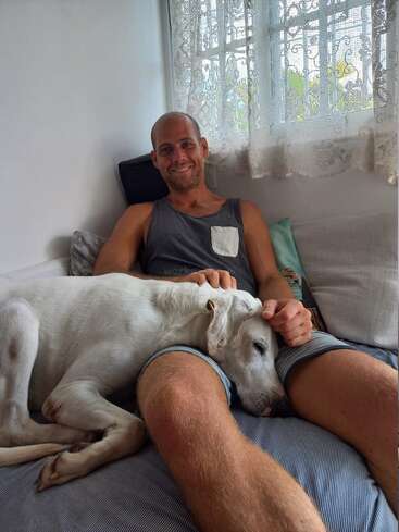Das Bild zeigt einen Mann, der auf einer Couch sitzt und einen weißen Hund auf seinem Schoß hat. Beide wirken entspannt und zufrieden in einer gemütlichen Umgebung.