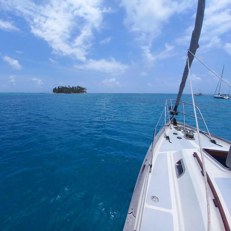 Das Bild zeigt eine ruhige Szene mit einem weißen Segelboot, das durch ruhige blaue Gewässer fährt, mit einer entfernten Insel am Horizont unter einem teilweise bewölkten Himmel.