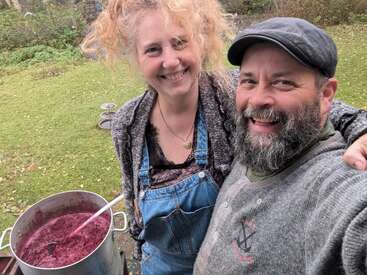 Um homem e uma mulher sorridentes posam ao ar livre, de braços dados. A mulher usa macacão e o homem, um boné. Um grande vaso de mistura roxa está próximo a você.