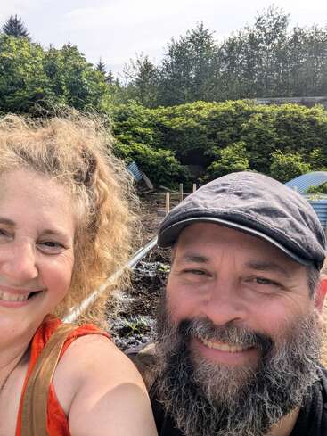 Um homem sorridente com barba e uma mulher com cabelos cacheados tiram uma selfie ao ar livre. Atrás deles, há vegetação, canteiros de jardim e a luz do sol de verão.